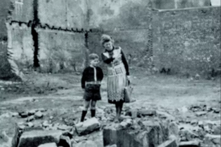 Ein Junge und eine Frau stehen zwischen Trümmern, verursacht durch Bombardierungen im Zweiten Weltkriegs.